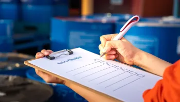 Safety inspection on the checklist document action with blurred background of chemical storage area at factory. Industrial safety concept photo. Selective focus at human's hand part.